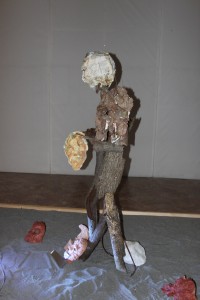 Il Guardiano: una figura antropomorfa fatta di corteccia d’albero, posta su uno strato di sabbia e circondata da tante maschere 